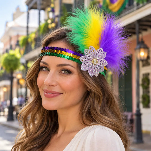 A woman wearing a festive Mardi Gras feather headband with green, yellow, and purple feathers, a purple flower embellishment, and a purple, gold, and green sequin band, photographed outdoors in a street setting.